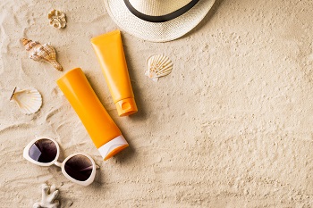 Tubes of sun tan lotion, sunglasses, and hat on a sandy beach with shells.